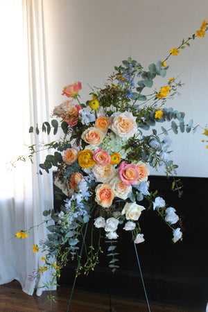 A floral standing spray arrangement with a variety of flowers including roses and other greenery, displayed on a easel arranged by Solabee