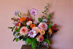 A vibrant floral arrangement featuring a variety of flowers in shades of pink, yellow, and white, with green foliage, displayed against a pink background.