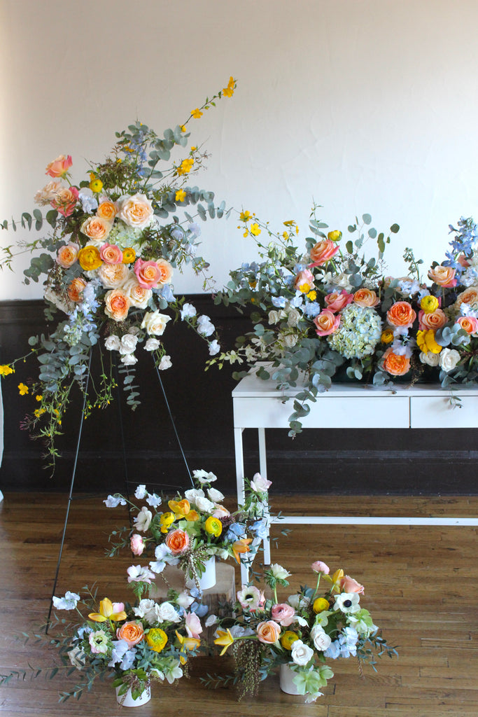 A funeral floral arrangement featuring a variety of flowers including white, yellow, and pink hues, displayed in a table setting with a black tripod and white backdrop.