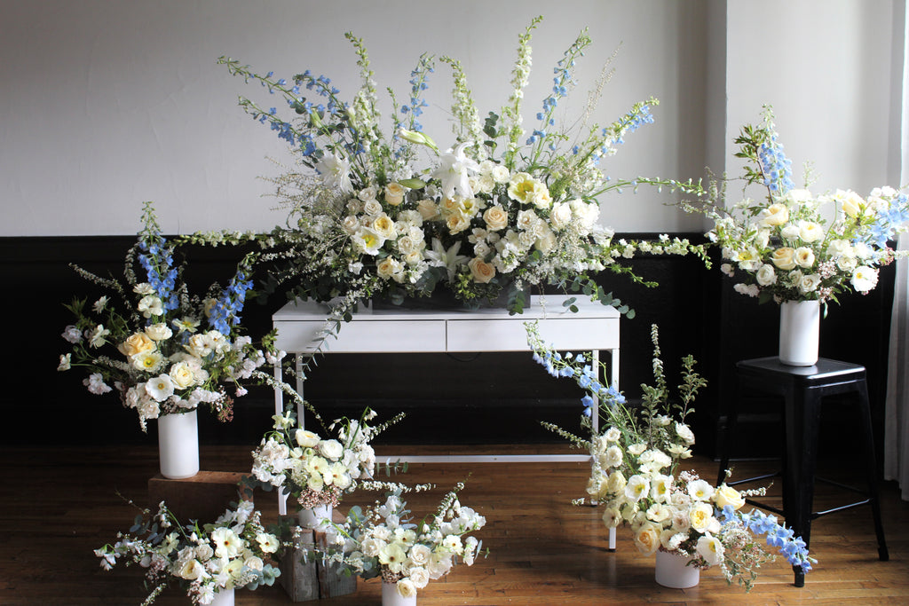 A series of floral arrangements for a funeral service, including a large arrangement on a white table, with multiple vases containing a mix of flowers predominantly in shades of white, blue, and green.