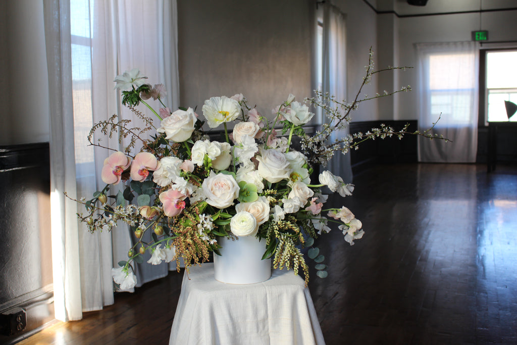 A floral arrangement with a variety of flowers in shades of white and pink, placed in a matte white vase, set against an indoor backdrop arranged by Solabee 
