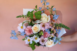 Bouquet of colorful flowers including roses, dahlias, zinnias, delphinium, and cosmos held against a pink background