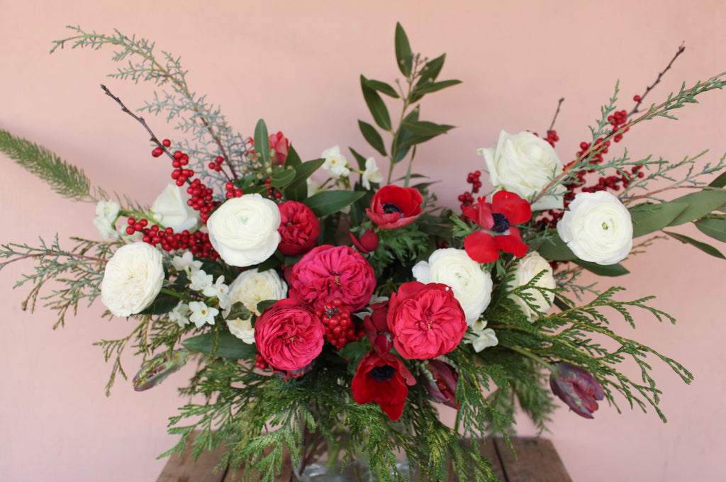 A holiday table centerpiece featuring a bouquet of winter flowers including red and white roses, greenery, and fragrant holiday greens.